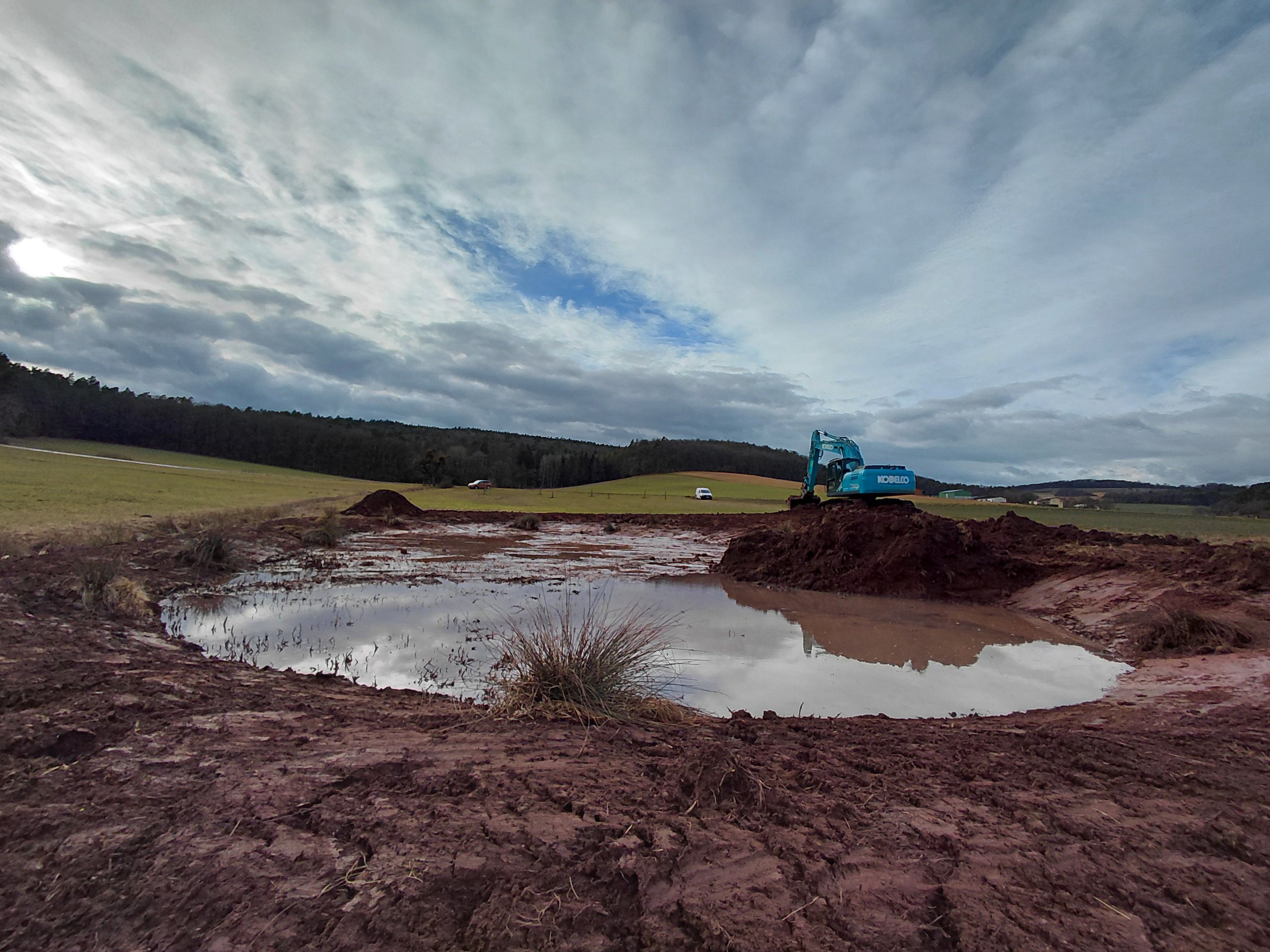 Frisch angelegte Wasserfläche in einer offenen Landschaft mit Feldern und Hügeln im Hintergrund