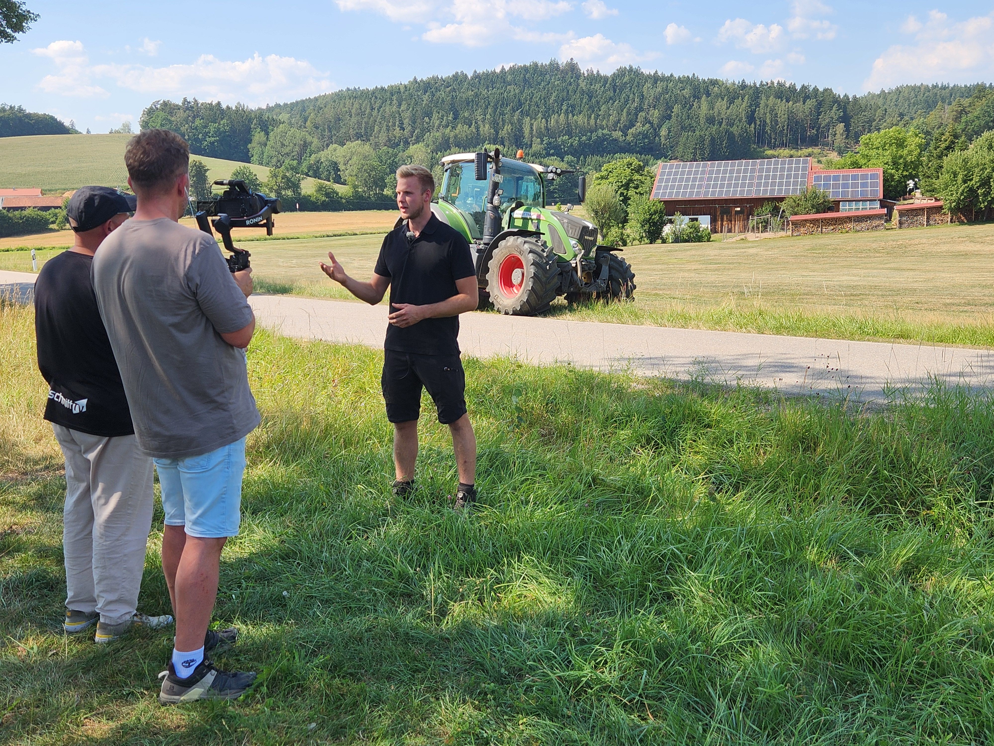 Drei Männer stehen auf einer Wiese, einer spricht gestikulierend, die beiden anderen filmen ihn, im Hintergrund ein Traktor