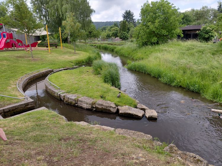 Tretbecken mit fließendem Wasser aus einem Fluss neben einem Spielplatz mit Klettergerüst und Rutsche.