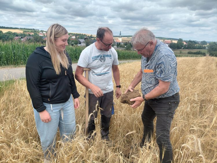Drei Personen stehen auf einem Getreidefeld, eine Person hält eine Bodenprobe in der Hand und zeigt sie den anderen beiden.