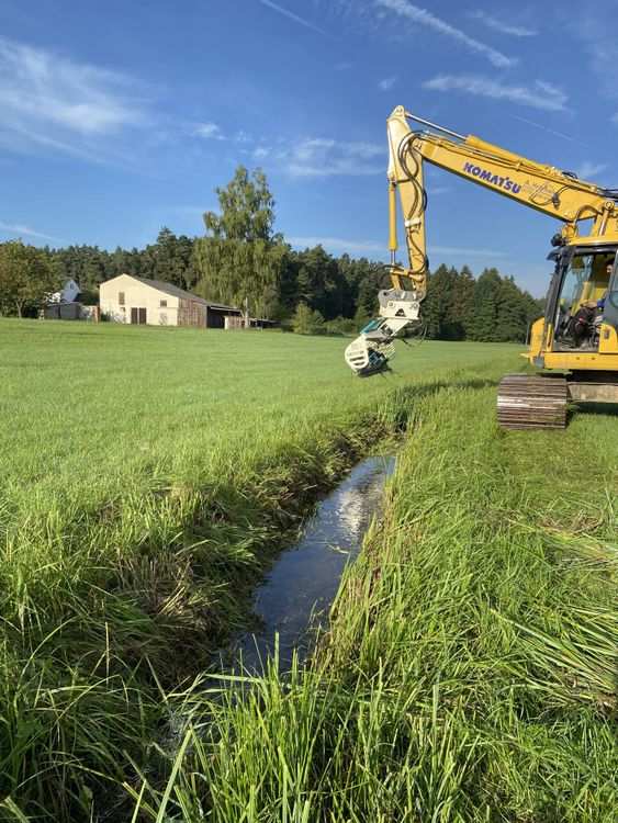 Bagger mit Mähkorb mäht Gras an einem Graben in einer Wiese vor Wald und Gebäuden.
