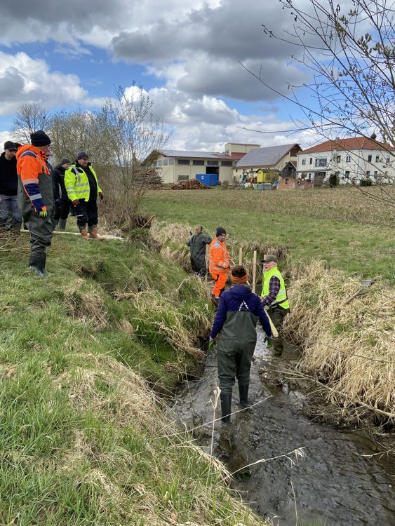 Mehrere Personen in Arbeitskleidung stehen an einem kleinen Bachlauf, einige stehen im Wasser, umgeben von Gras und Büschen, im Hintergrund Häuser und bewölkter Himmel